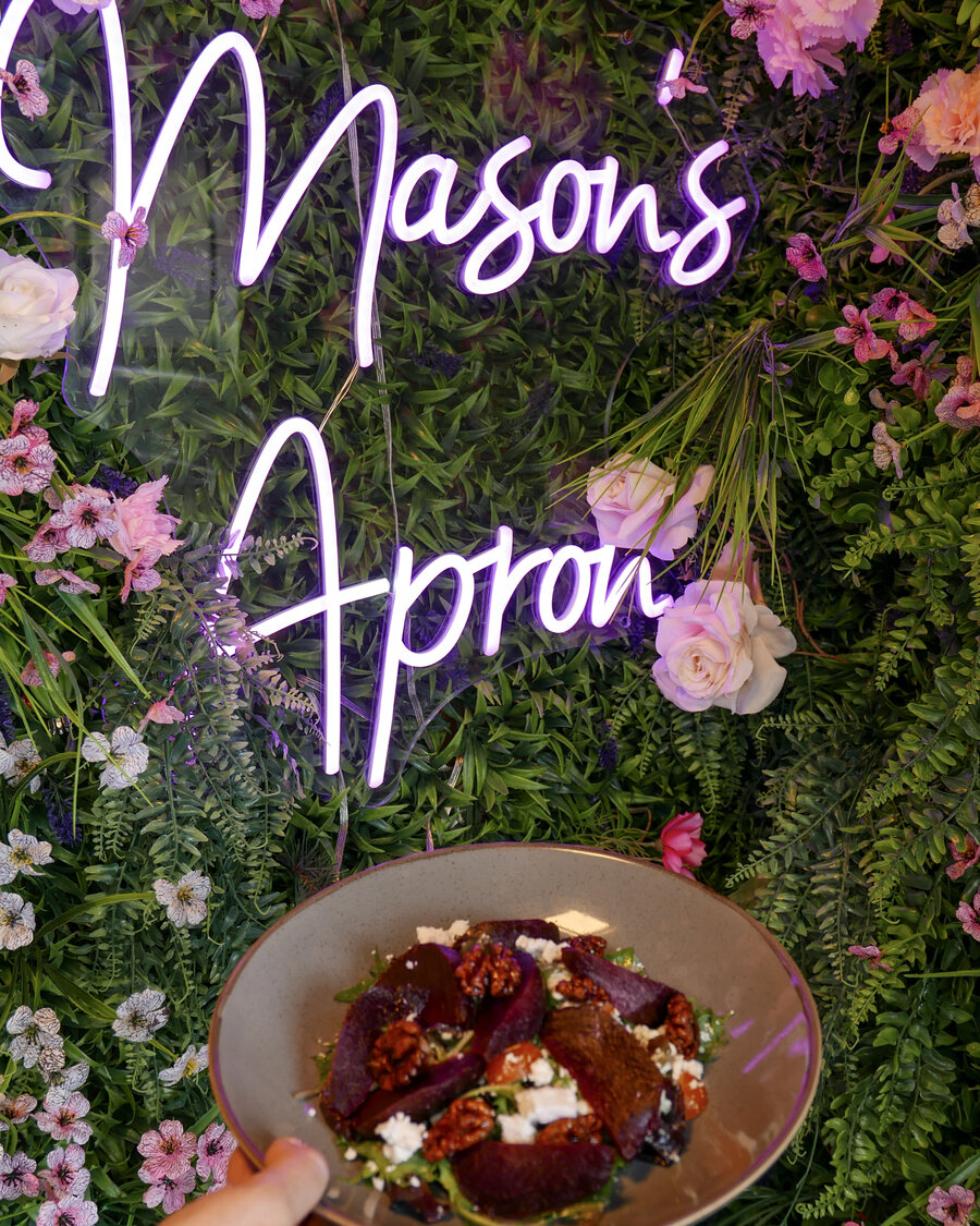 The Mason's Apron neon sign on a flower wall